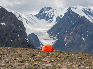 Summer camping in mountain. Bright alpine landscape with vivid orange tent at very high altitude with view to high mountain and large glacier in dramatic clouds. Awesome mountain scenery with tent.