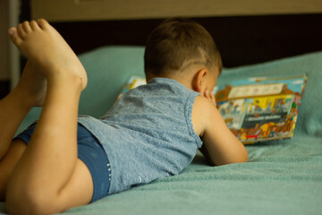 Little, cheerful, three years old boy lying on his belly on the sofa, with open book in front of him.