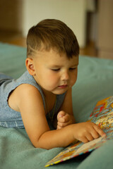 A cute little boy with broun hair and a blue shirt is lying on the bed reading a book