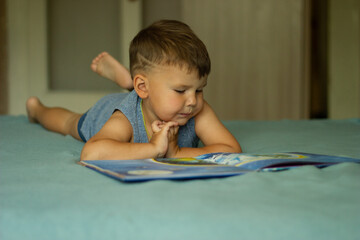 Dressed in casual clothing cheerful boy reads book lying down on a bed in living room.