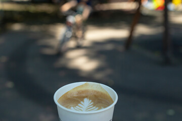 Shallow depth of field photography of a cappuccino paper cup , with a blurred bicycle in a park. 