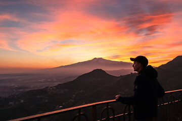 Fototapeta premium Tourist man watching beautiful sunset behind volcano Mount Etna near Castelmola, Taormina, Sicily, Italy, Europe, EU. Clouds with vibrant red orange colors. Silhouette of person during twilight. Awe