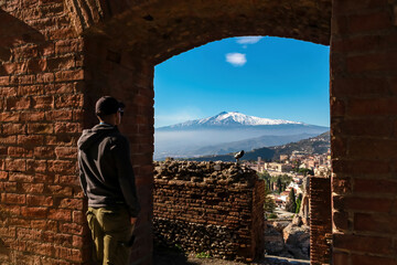 Tourist man in hat with panoramic view on snow capped Mount Etna volcano on a sunny day seen from...