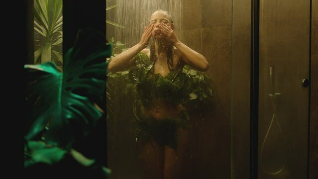 Beautiful latin brunette woman taking a shower , wearing leaf swimwear . Bathroom designed with plants , trees tropic concept . Black tropical girl showering inside modern bathroom . Slow motion