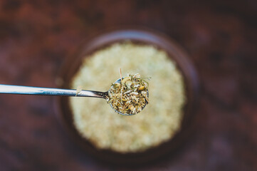 Full spoonful of dried Matricaria chamomilla flowers above the kitchen table. Brown bowl of medicinal herbs blurred in the background. Process of making an herbal tea or infusion of chamomile.