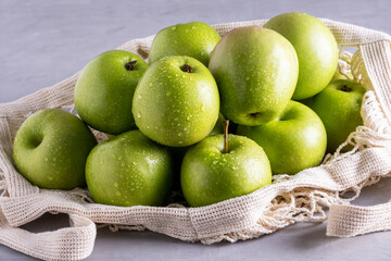 Several fresh ripe green apples lie in an eco-friendly bag on a gray surface. Selective focus.