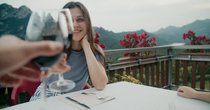 Happy woman clink glasses of red wine with boyfriend sitting in restaurant with beautiful mountain views at sunset. Traveler couple testing alcoholic drink on summertime vacation journey.
