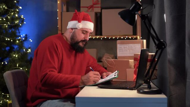 Dropshipping owner male Hindu Santa Claus works at home office at night on laptop, checks order for confirmation before shipping to customer during Christmas sale. Online store and small business