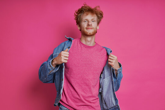 Handsome redhead man in denim clothing standing against pink background