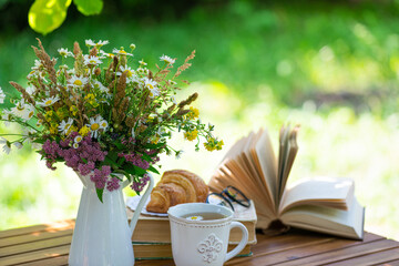 Bouquet of meadow flowers, croissant, cup of tea or coffee, books on table in summer garden. Rest in garden, reading books, breakfast, vacations in nature concept. Summertime in garden on backyard