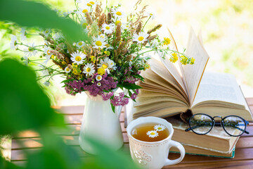 Bouquet of meadow flowers, croissant, cup of tea or coffee, books on table in summer garden. Rest...