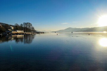 Obraz premium View on Woerthersee from Poertschach in Carinthia, Austria. Calm lake reflecting the landscape. View on the Karawanks Alps and Pyramidenkogel. Sunset sunrise vibes. Villages at the waters edge. Reed