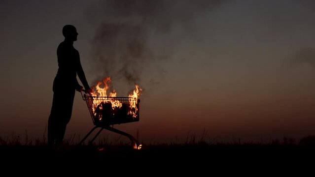 A man rolls a burning cart from a supermarket in an empty field, against the background of a golden sunset closer to the dark night, slow motion