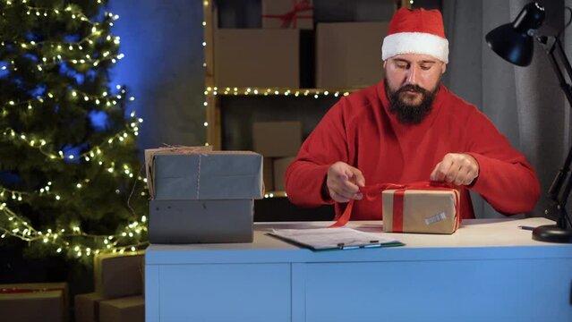 Dropshipping owner male Hindu Santa Claus works at night in his home office, knitting a big red bow on box before shipping to a customer during the Christmas sale. Online store and small business.