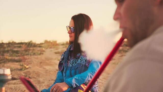 People smoke hookah on sandy beach. Focus shifts from beautiful girl with dark hair to the guy exhaling thick clouds of smoke against the background of the setting sun in slow motion. Summer. Vacation