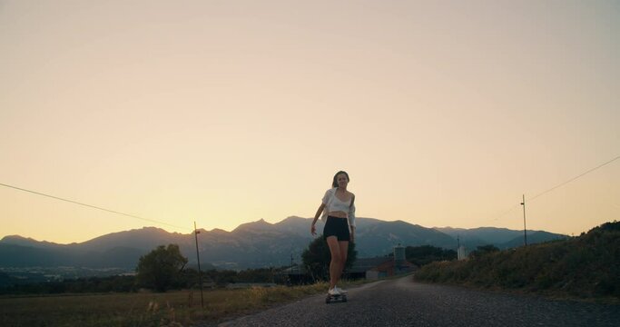 Excited young adult woman ride on skate longboard and successfuly rise her hands on sunset with mountain view. Concept of youth and freedom active lifestyle outdoors. Happy girl skateboarding on road
