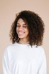 Portrait of smiling African american girl winking one eye mouth, open laugh laughter wearing casual white t-shirt isolated beige background    