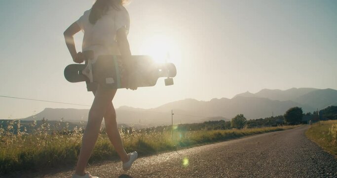 Skater girl holding skate longboard and walk along meadow on country road. Concept of youth and active lifestyle outdoors. Active happy forever young woman on summertime vacations with mountain view