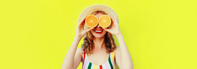 Summer portrait of happy cheerful smiling woman covering her eyes with slices of orange and looking for something wearing straw hat on yellow background