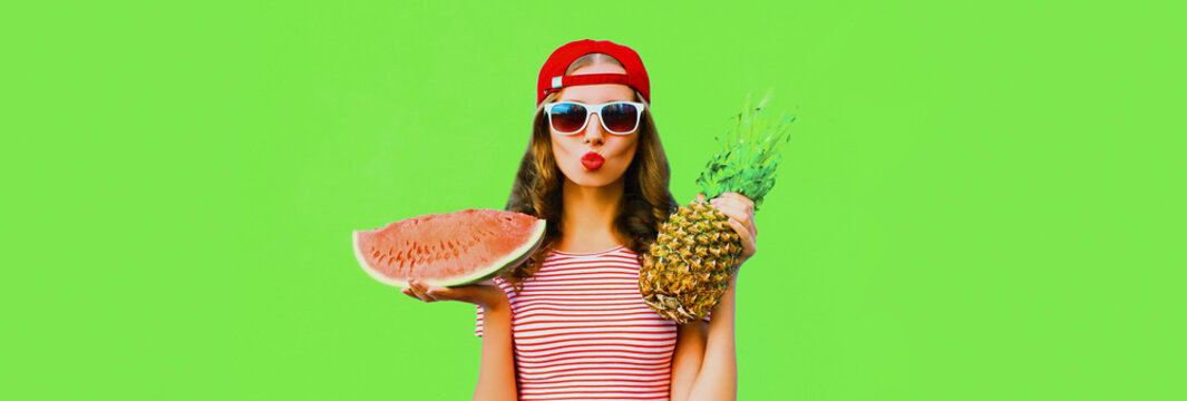 Portrait of happy young woman model blowing her lips posing with pineapple and slice of watermelon on green background, blank copy space for advertising text
