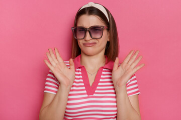 Portrait of Caucasian confused woman wearing striped T-shirt and sunglasses posing isolated over pink background, raised arms, looking at something unpleasant. - obrazy, fototapety, plakaty