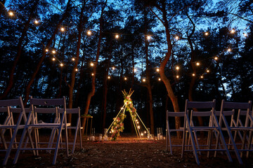 Festive string lights illumination on boho tipi arch decor on outdoor wedding ceremony venue in...