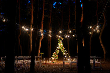 Festive string lights illumination on boho tipi arch decor on outdoor wedding ceremony venue in...