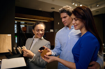 Cheerful mature couple testing wood surfaces for kitchen countertop in furniture design showroom while standing next to pleasant interior designer presenting many different samples of surfaces