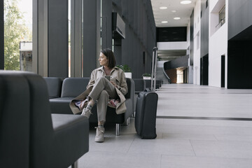 woman at the airport or congress center with a suitcase is sitting in the waiting area and chatting on her smartphone. a beautiful European woman in a beige trench coat at the airport is waiting to