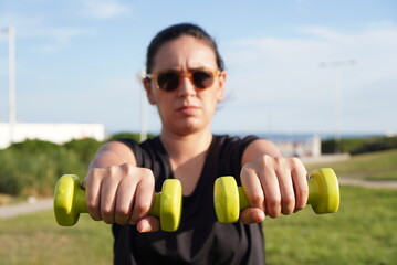 Young and attractive woman does sports in a park near the beach lifting weights with her arms, she dresses in dark clothes and wears glasses, concept of new challenges 2022
