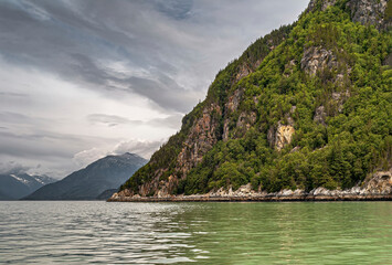 Obraz premium Skagway, Alaska, USA - July 20, 2011: Taiya Inlet above Chilkoot Inlet. Wide fjord landscape with green forested mountain flank rising out of green ocean water. Mountains on horizon under gray cloudsc