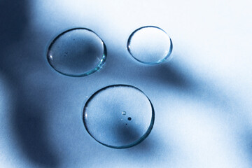 Transparent drops of a cosmetic product on a blue background with an abstract shadow. Hyaluronic acid, peptides, body care serum.