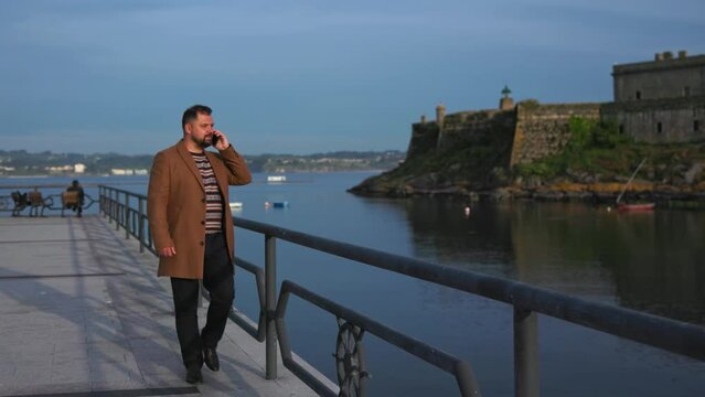 A business man with a full-length beard in a brown coat walks along the embankment with a phone. A man in dark blue pants, classic brown shoes and a patterned sweater. A man walks along a pier near a