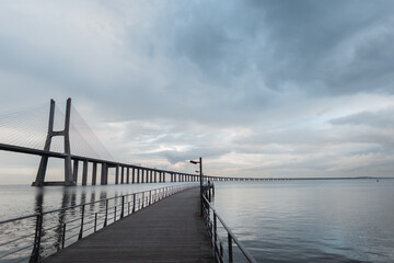 Fototapeta na wymiar Amazing Long Bridge Vasque da Gama with wooden pier, sea and clouds in Lisbon, Portugal.