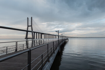 Fototapeta na wymiar Amazing bridge and wooden pier on a gray sky with clouds. Lisbon, Portugal