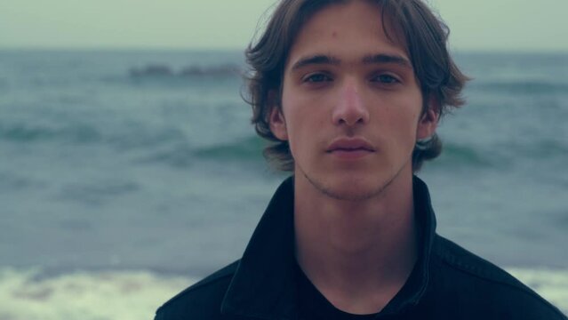 Young man  on the empty ocean beach, on the cloudy rainy weather. Confident and calm guy is on a sandy shore looks at the camera. Closeup face of handsome man posing outdoors. Slow motion 4k footage.
