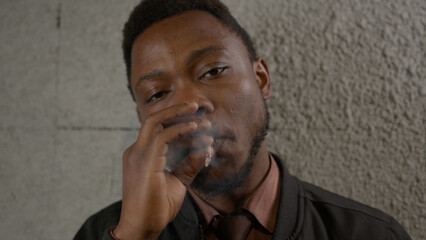 Fototapeta na wymiar Nicotine addiction in black skin man standing near wall and smoking cigarette exhaling smoke. Portrait African-American man puts cigarette to his mouth and inhales smoke into his lungs and exhales.