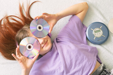 Happy teenage girl in lilac t-shirt lies on bed in pink headphones holds cd disks in her hands