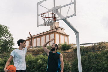 Happy young boys shaking hands after a street basketball game. Front view image of a couple of sporty men with an orange ball with their hands raised to join them. Concept of friendship and sport