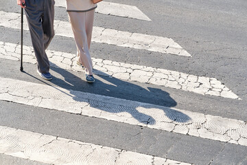 two unrecognizable elderly people, one of them with a cane, crossing a pedestrian crossing