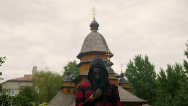 Young religious caucasian girl in hood performing the sign of the cross with old wooden christian church in the background. Beautiful hooded woman confessing sins to God and acknowledges her mistakes 