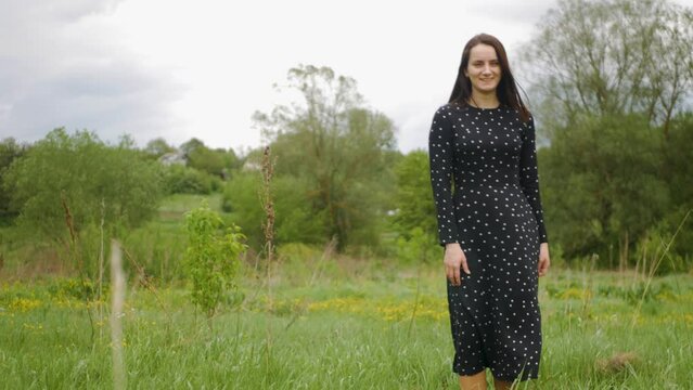 Attractive white woman in black summer dress spinning and smiling in middle of beautiful green meadow. Long-haired caucasian model posing in grass-covered field outdoors. Charming female in nature
