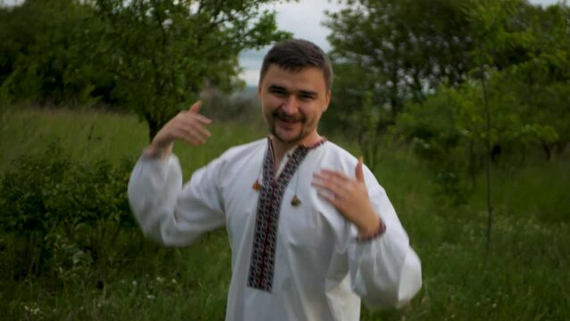 Cheerful guy in traditional Ukrainian national outfit dancing and swinging arms around. Happy adult male in white embroidered vyshyvanka smiling and twirling while celebrating victory and success
