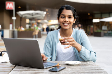 Portrait of smiling Indian woman freelancer using laptop drinking coffee sitting in cafe. Coffee break