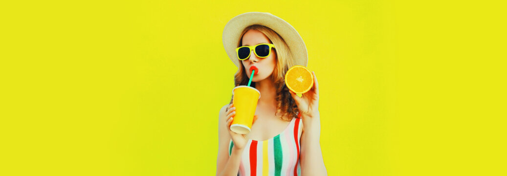 Summer portrait of beautiful young woman drinking fresh juice with slice of orange fruits wearing straw hat, sunglasses on yellow background