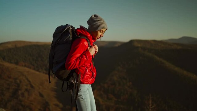 slow motion view from behind adventurous female hiker backpacker in red jacket with backpack stands on mountain top at sunset above the woods. freedom and solo traveling concept. unity with nature.