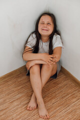  portrait of little scared girl sitting with knees up, crossed arms, in corner on floor, covering face. Protest against domestic violence, abuse, fighting, conflict, bullying. Vertical.