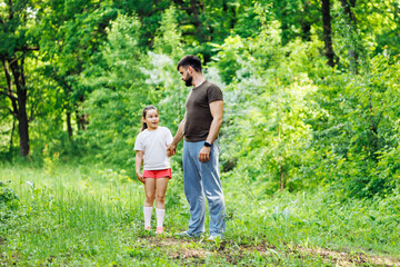 Naklejka na ściany i meble Portrait of wonderful family walking in forest around green trees. Little daughter holding hand of middle-aged bearded father. Surprised girl looking at man attentively. Summer activities, vacation.