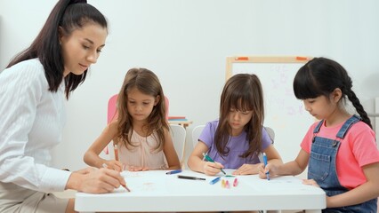 Fototapeta premium Woman teacher teaching little girl to paint color book on the table in classroom,kindergarten education school.Multi-ethnic preschool teacher and students in classroom.Kindergarten and study concept.