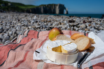 Lunch with french round camembert cow cheese from Calvados region, fresh baked baguette bread and apple on pebbles stones beach with view on alebaster cliffs Porte d'Aval in Etretat, Normandy, France